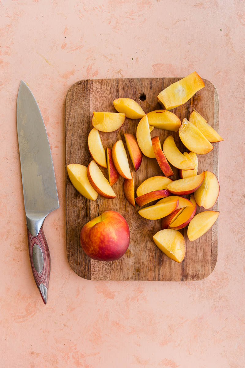 Knife and peach slices on a cutting board with a large knife on the side.