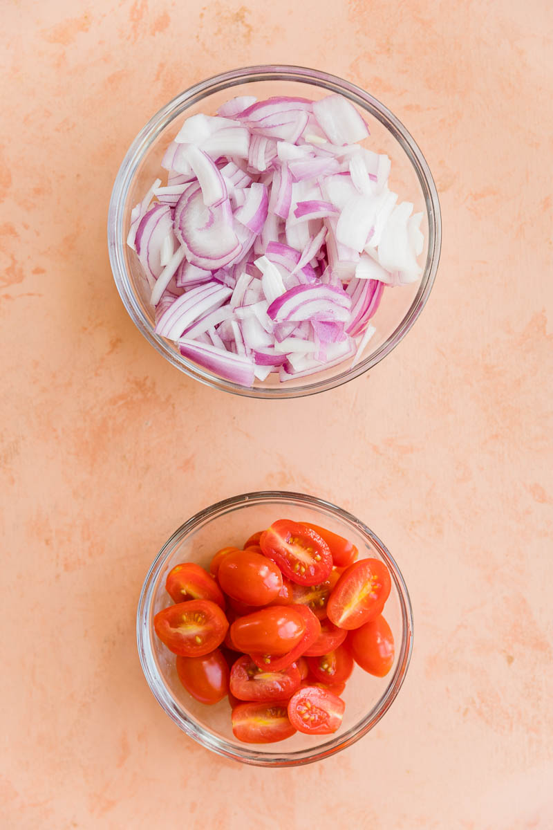 Thinly sliced red onions in a bowl and grape tomatoes cut in half in a bowl.