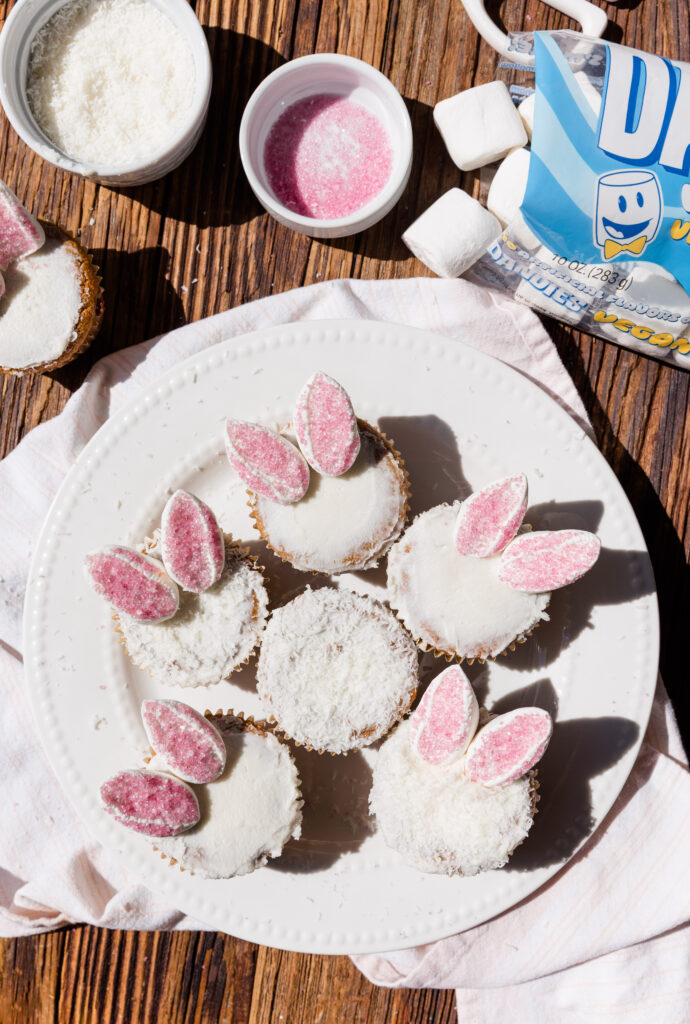 Overhead picture of frosted cupcakes dipped in shredded coconut and topped with pink marshmallow bunny ears.