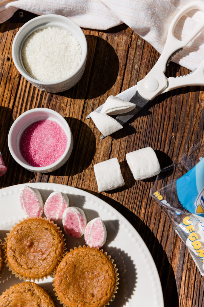 Scissors cutting marshmallows in half with pink sprinkles and cupcakes on the side.