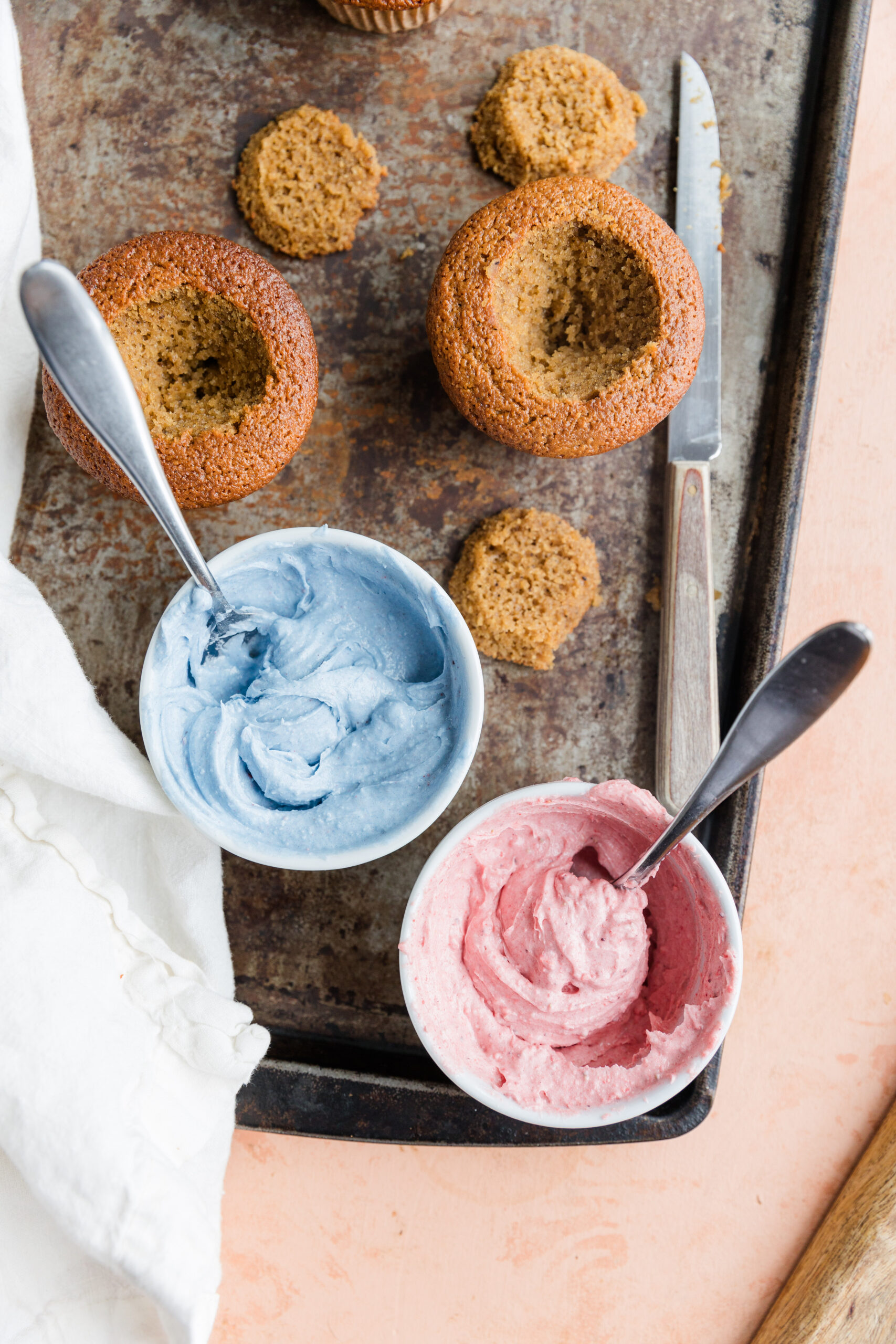 Vanilla cupcakes with a hole cut in the center next to a bowl of blue frosting and pink frosting.